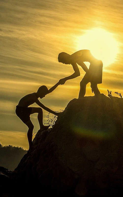 silhouette of boy helping another boy climb up to the top of a steep hill bright sun in behind